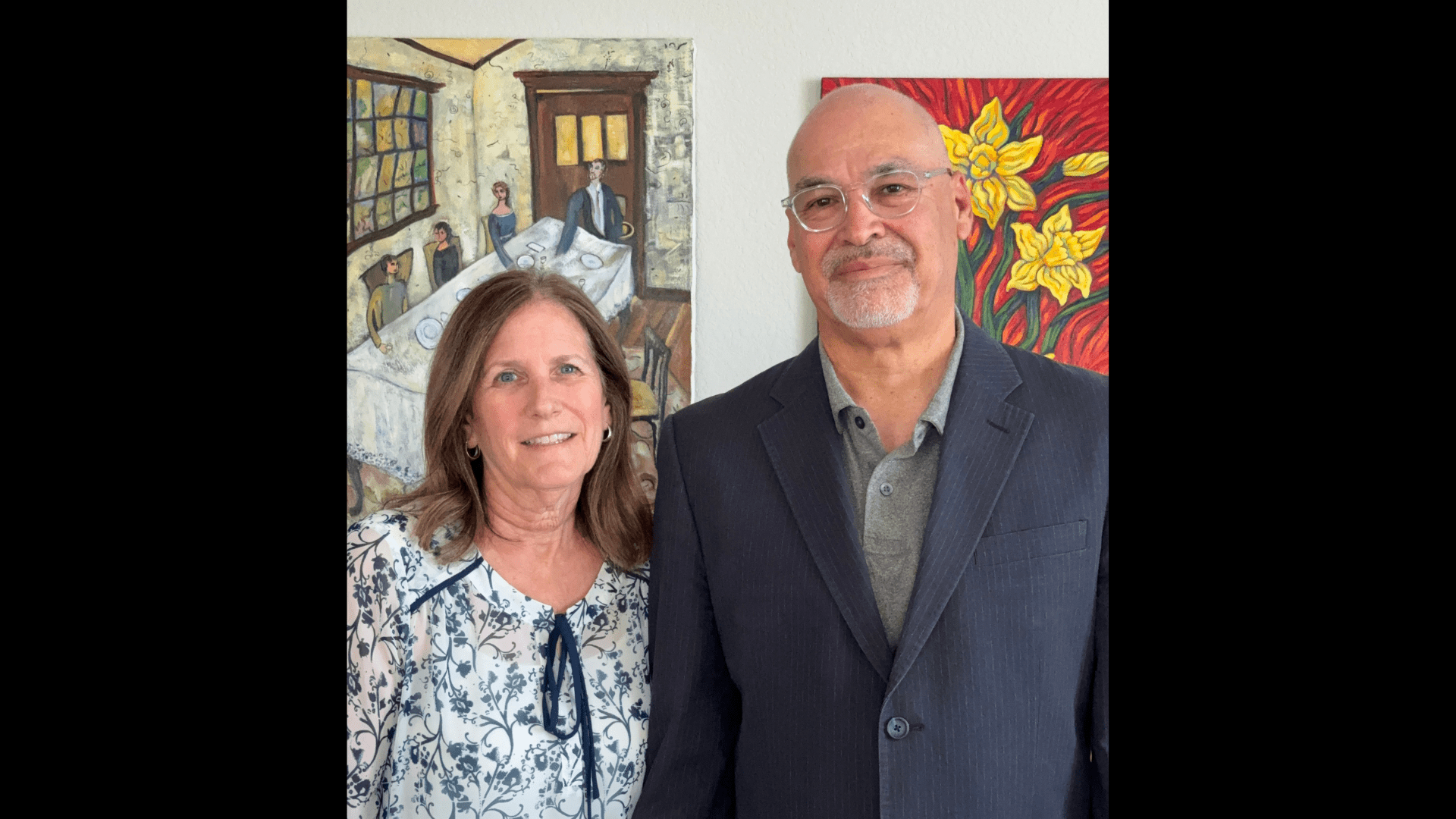 Jo Ann and Michael Montoya pose for a photo, a wall with paintings behind them.