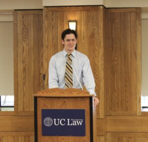 Alex Nicholson, wearing a long-sleeved shirt, tie, and eyeglasses, speaks at a podium with UC Law SF's logo on it.