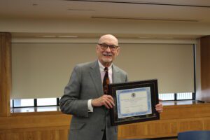 Professor Matt Coles holding his award in front of a closed window wearing a suit and tie.