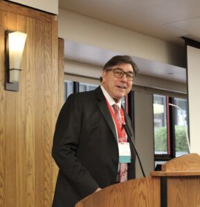 David Faigman stands at a podium in front of a microphone, wearing eyeglasses, a dark suit jacket, and tie.