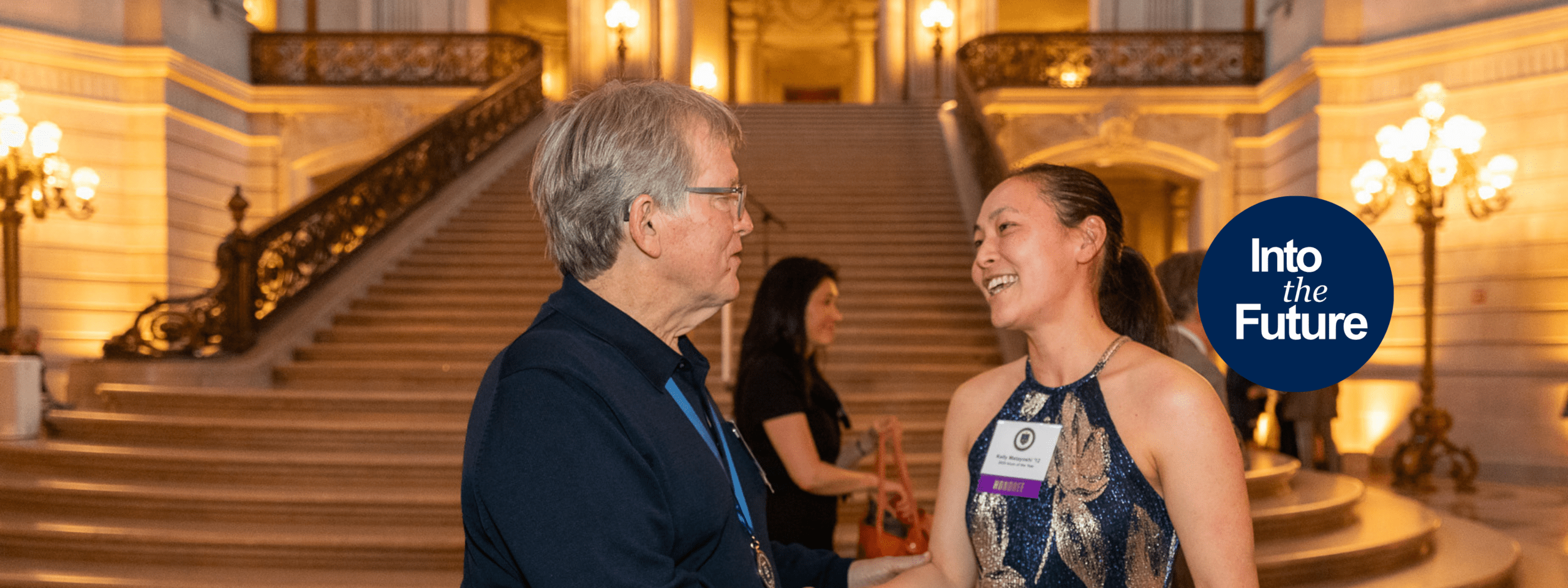 Two UC Law SF alumni talking to each other at the 2025 Spring Week All Alumni Party held in San Francisco's City Hall. On the right side of the image is the Into the Future campaign logo