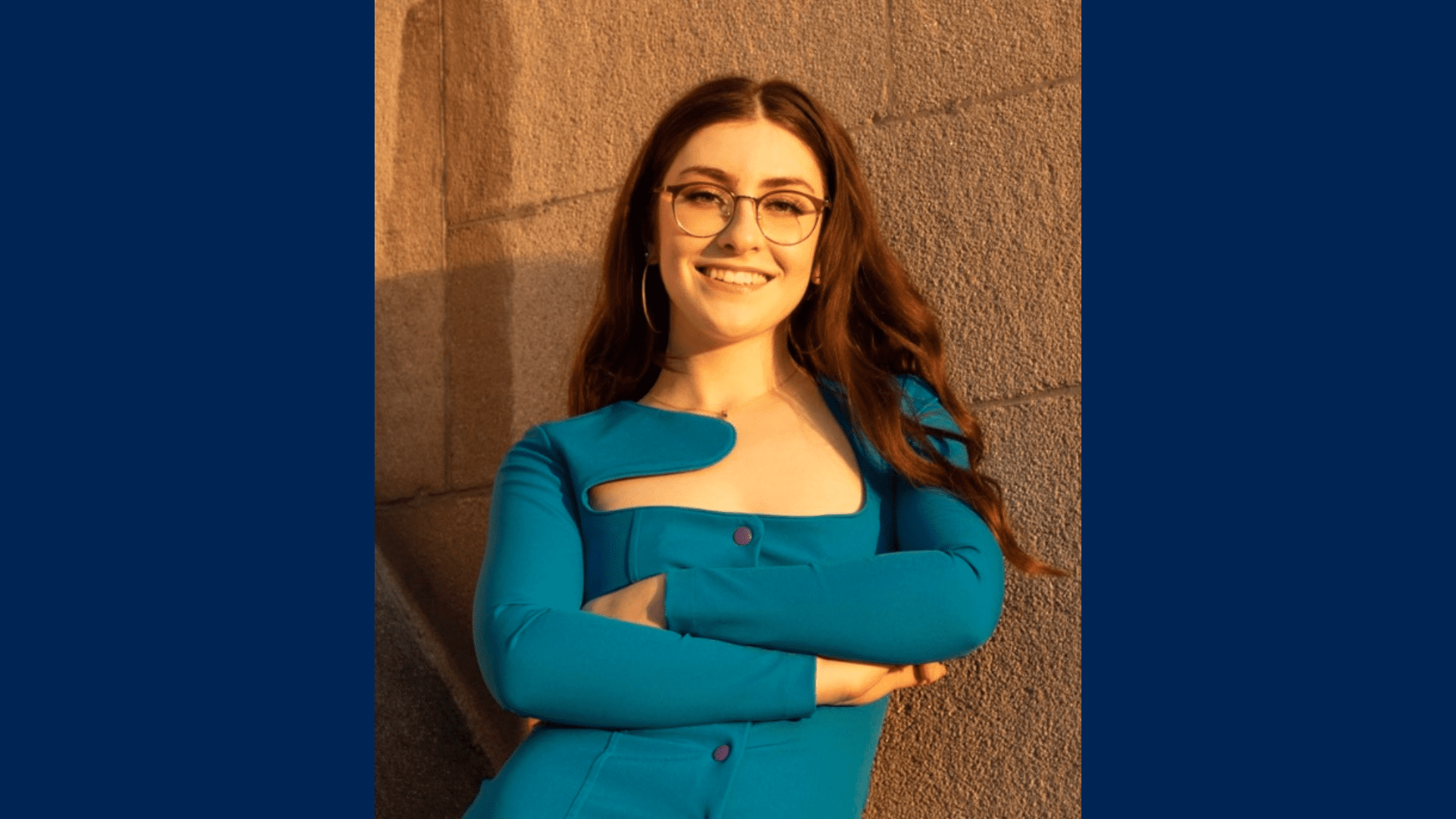 3L Emily Cole wearing a blue shirt and glasses in front of a brick wall.