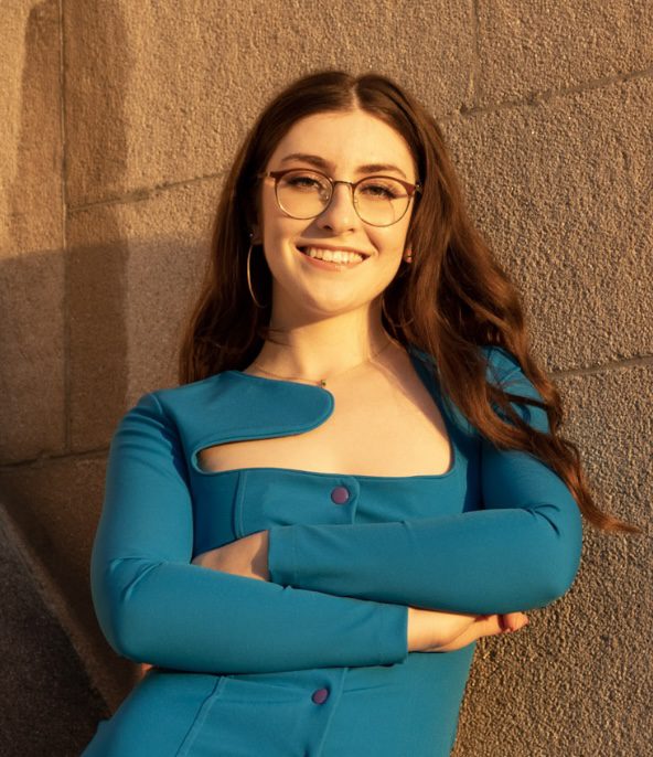 3L Emily Cole wearing a blue shirt and glasses in front of a brick wall.