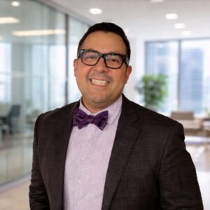 3L Juan Herrera wearing a suit, bowtie, and glasses in an office.