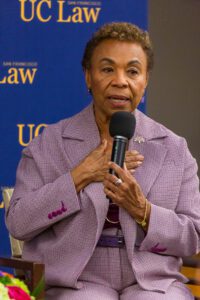 Barbara Lee speaks into a microphone, holding her hand on her chest.