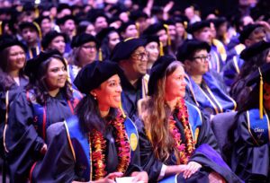 Seated graduates smile at the front of a crowd of people in caps and gowns.