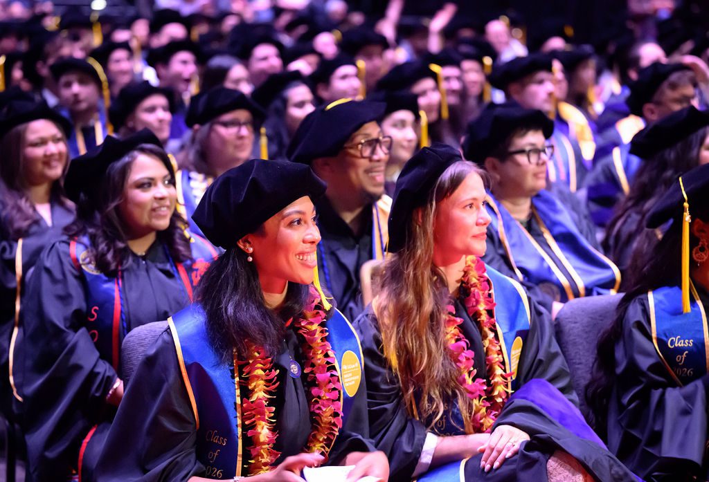 Seated graduates smile at the front of a crowd of people in caps and gowns.