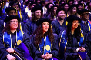 Three seated graduates smile with a crowd of seated graduates behind them.