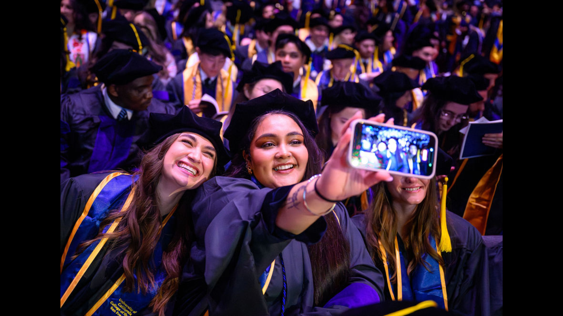 A graduate holds a phone in front of her to take a selfie with a fellow graduate, a crowd of graduates behind them.