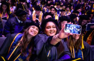 A graduate holds a phone in front of her to take a selfie with a fellow graduate, a crowd of graduates behind them.