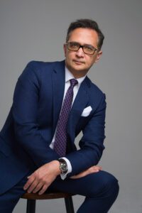 a headshot photo of Farzad Tabatabai in a blue suit and purple tie wearing eyeglasses.
