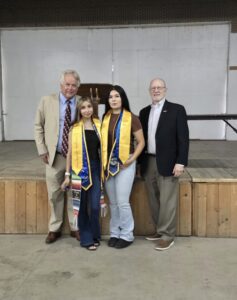 Four people posing for a photo. The two high school students in the center wear graduation stoles