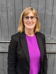 Melissa Hooper wears eyeglasses and smiles while posing in a pink shirt and black suit jacket in front of a gray wooden fence.
