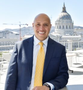 Mario Ernesto Lopez poses in a blue suit and yellow tie with the San Francisco City Hall dome behind him.