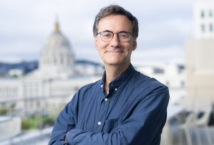 Reuel Schiller poses in a blue button-up shirt with San Francisco City Hall in the background.